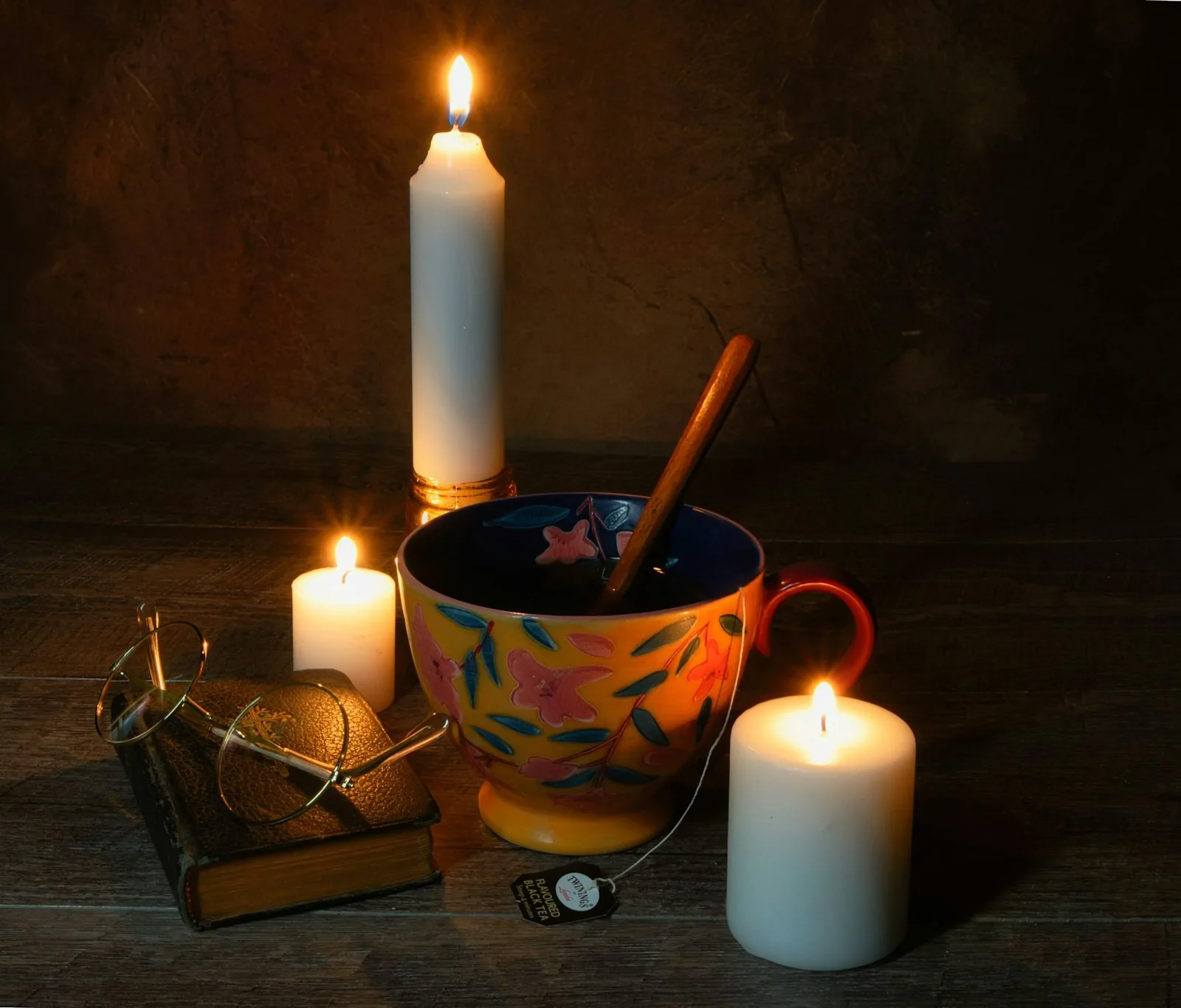 A colorful teacup beside candles, a small book and reading glasses